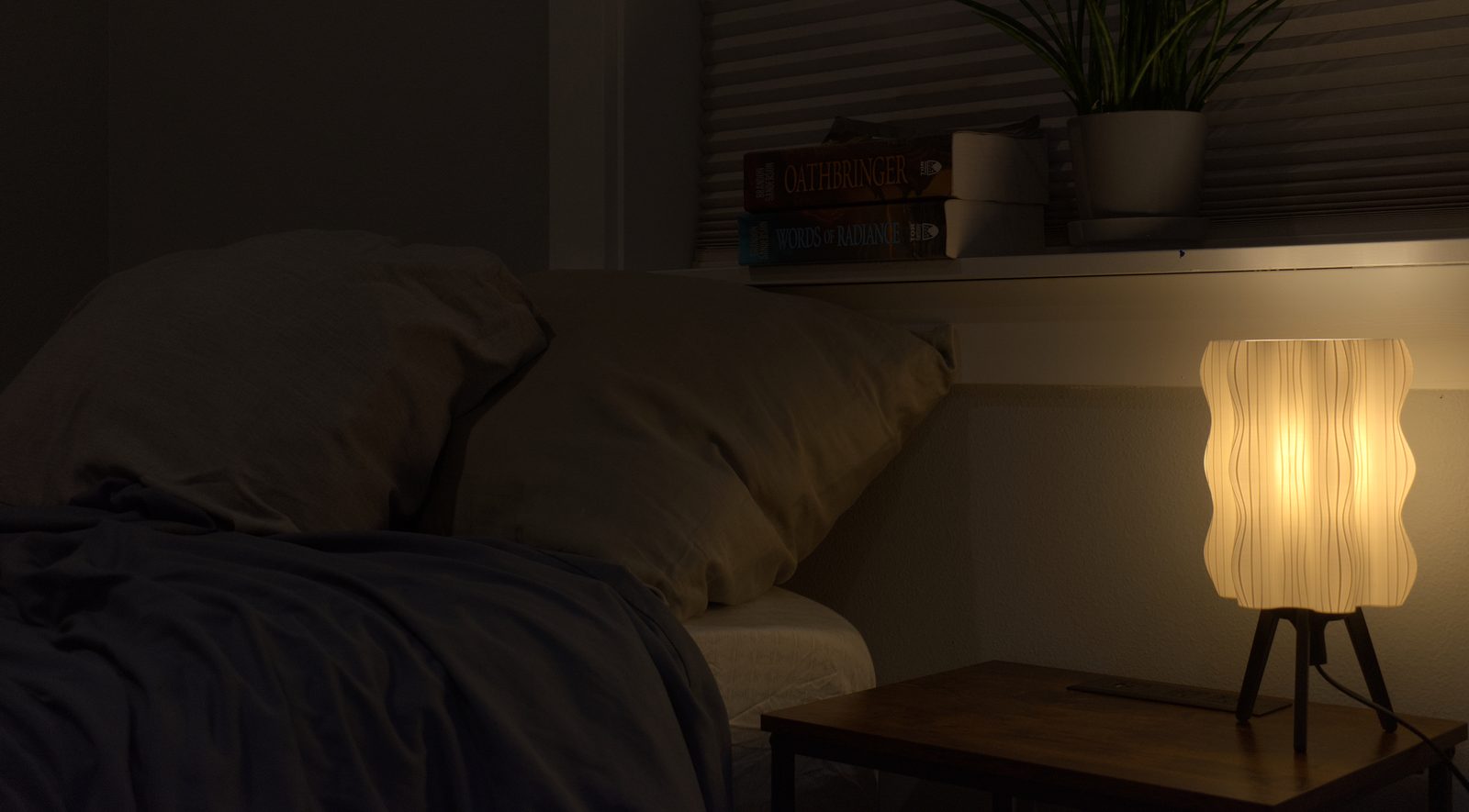 A white Ripple Lamp glowing on a bedside table in a cozy dark bedroom